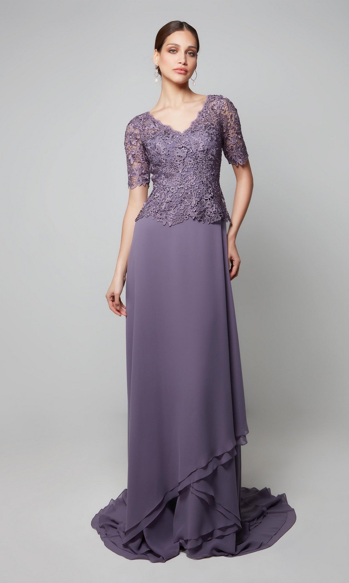 Wearing the Alyce 27556, a long formal lavender dress with a beaded lace bodice and short sleeves, a woman stands against a plain gray background—an elegant pick for any mother-of-the-bride.