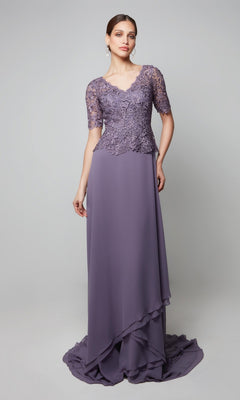 Wearing the Alyce 27556, a long formal lavender dress with a beaded lace bodice and short sleeves, a woman stands against a plain gray background—an elegant pick for any mother-of-the-bride.