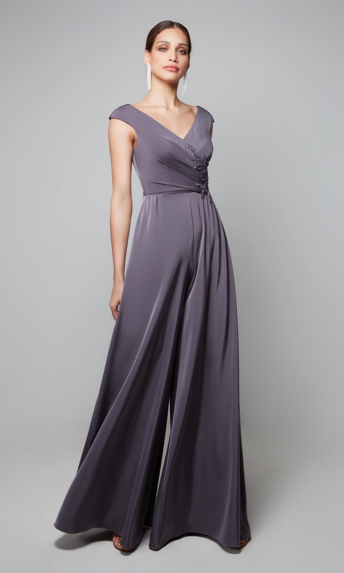 A woman wears the Alyce 27559 long formal jumpsuit in graphite, featuring cap sleeves, a V-neckline, and decorative waist detail, standing against a plain background.