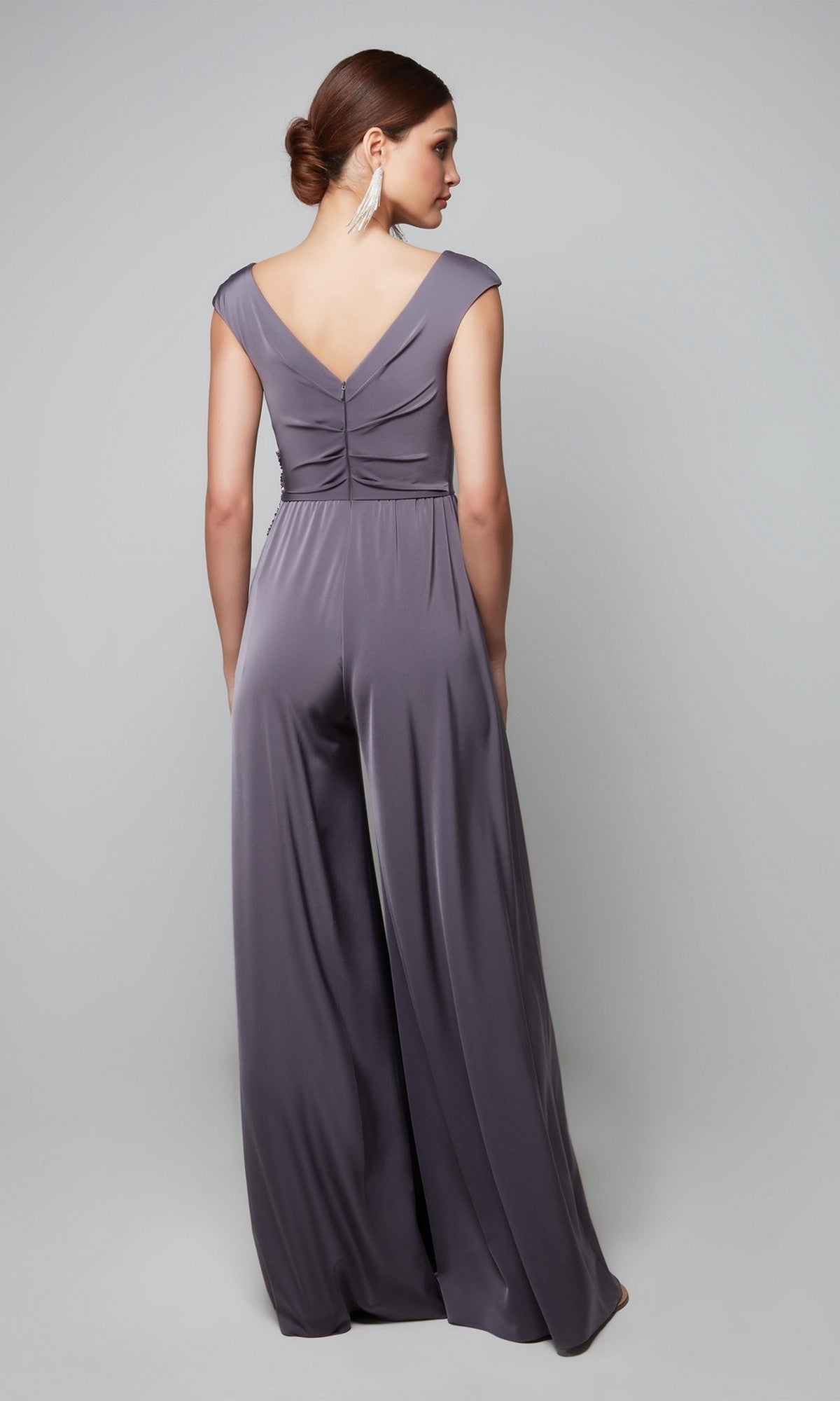 A woman stands with her back to the camera in a silky lavender-gray Alyce 27559 long formal jumpsuit, featuring a sleeveless design, deep V-back, and wide legs. She wears her hair in a low bun and pairs it with long earrings.