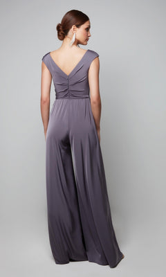 A woman stands with her back to the camera in a silky lavender-gray Alyce 27559 long formal jumpsuit, featuring a sleeveless design, deep V-back, and wide legs. She wears her hair in a low bun and pairs it with long earrings.