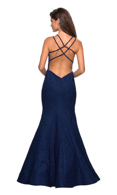 A woman wearing the Backless Lace Mermaid Prom Dress: La Femme 27560—a navy blue, floor-length, fitted gown with a flared hem and crisscrossed open-back straps—looks stunning and ready to make an entrance at prom.