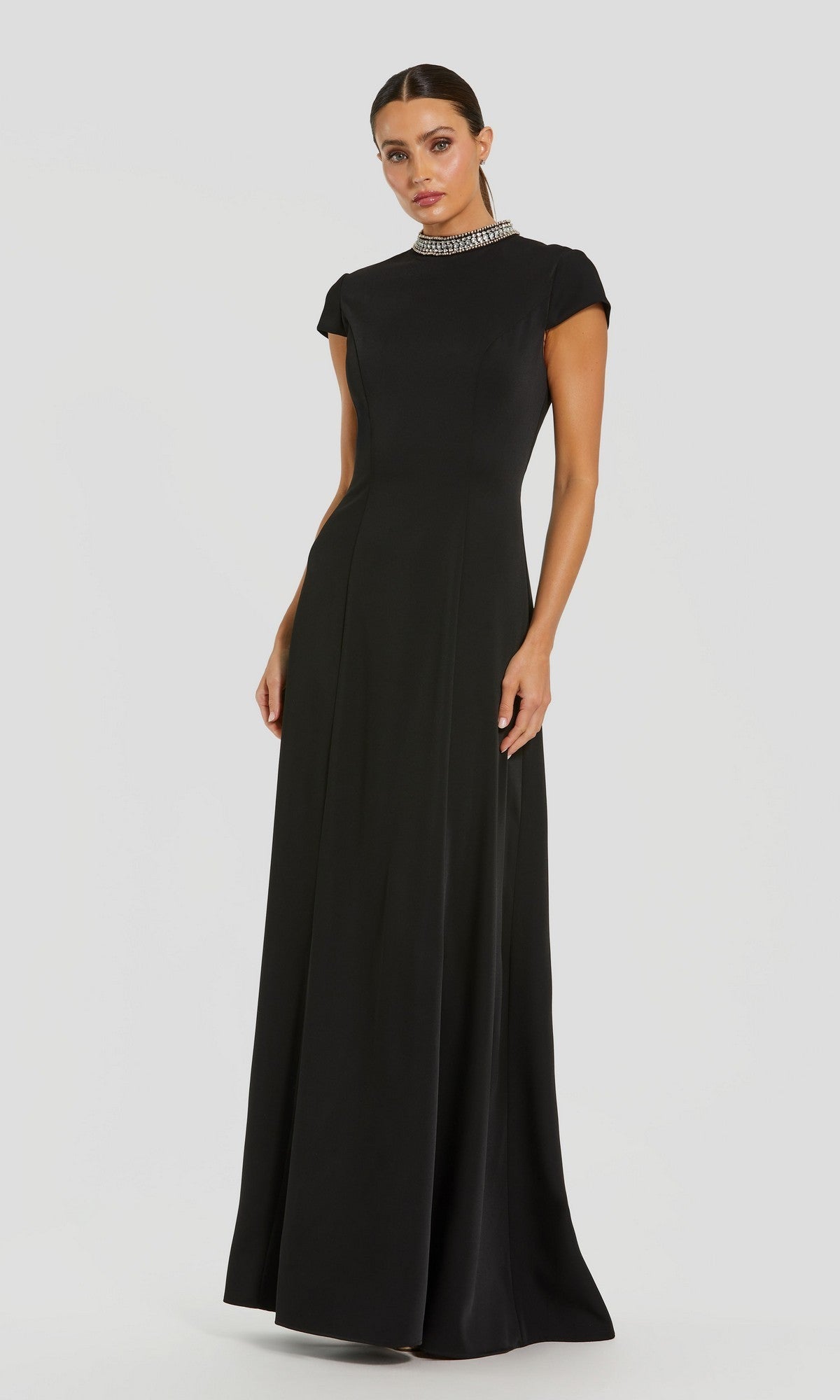 A woman wearing the Mac Duggal 27564 long formal black dress stands against a plain light background. This floor-length crepe gown features short sleeves, a high neckline, and a jeweled collar, capturing timeless sophistication in every detail.
