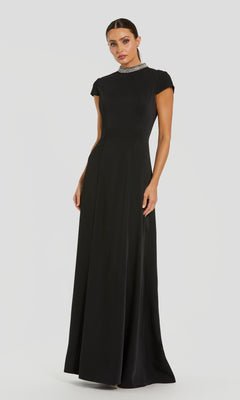 A woman wearing the Mac Duggal 27564 long formal black dress stands against a plain light background. This floor-length crepe gown features short sleeves, a high neckline, and a jeweled collar, capturing timeless sophistication in every detail.