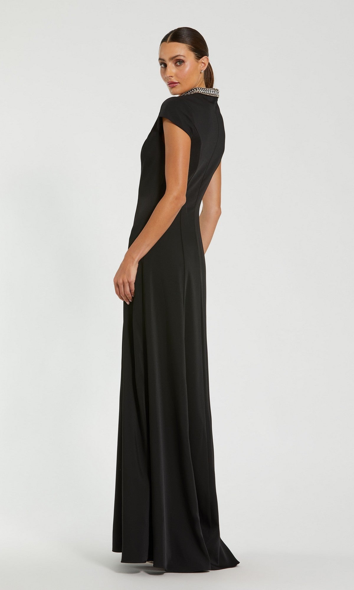A woman wearing the Mac Duggal 27564 long formal dress—a floor-length black gown with short sleeves and an embellished collar—stands against a plain background, looking over her shoulder.