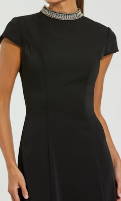 A woman is shown from shoulder to thigh against a plain light background wearing the Mac Duggal 27564 long formal dress, which features a fitted silhouette, short sleeves, and a jeweled collar in classic black.