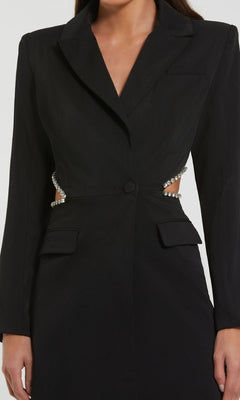 A woman wears the Mac Duggal 27520 Short Cut-Out Black Party Dress— a tailored blazer dress with long sleeves, cutout sides, rhinestone embellishments, and flap pockets, making it a chic cocktail choice.