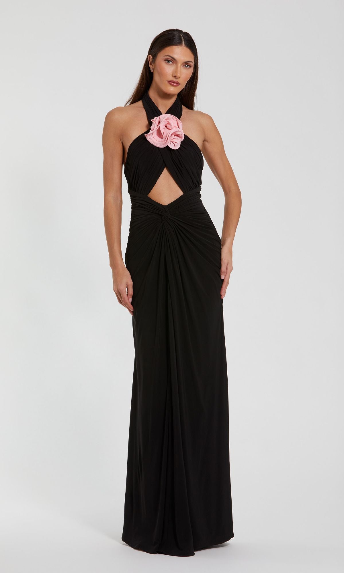 A woman wears the Mac Duggal 27572 Cut-Out Black Halter Prom Dress, a formal sleeveless gown featuring a front cutout, gathered waist, and a large pink flower detail at the neckline.