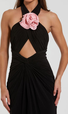 A woman wears the Cut-Out Black Halter Prom Dress, Mac Duggal 27572—a striking black halter gown with a central cutout and a bold pink flower at the neckline, ideal for formal evening events.