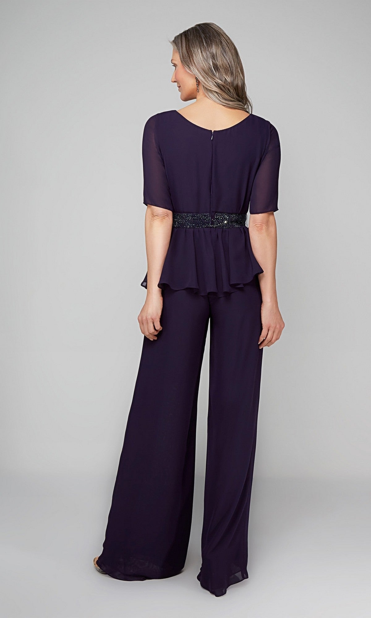Woman stands with her back to the camera, wearing the Alyce 27576 Long Formal Jumpsuit—dark purple with wide-leg pants, a peplum top, and beaded waistband—set against a plain light gray background.