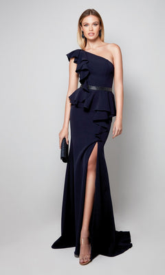 A woman in a midnight Alyce 27577 long formal dress—one-shoulder with ruffle detail, beaded belt, thigh-high slit—paired with clear heels and a black clutch, stands against a plain background.