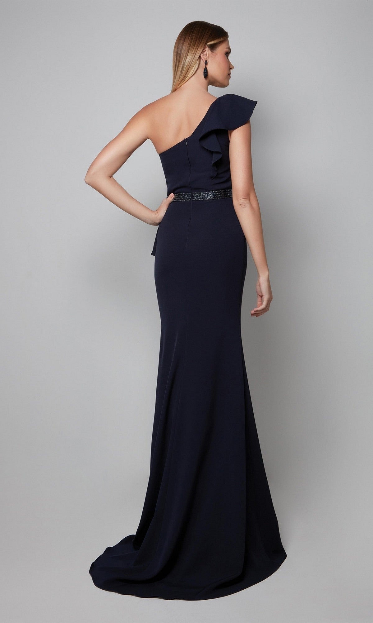 A woman wearing the Long Formal Dress: Alyce 27577—a navy blue, one-shoulder gown with a fitted bodice, ruffle detail, and floor-length skirt—stands against a plain gray background.