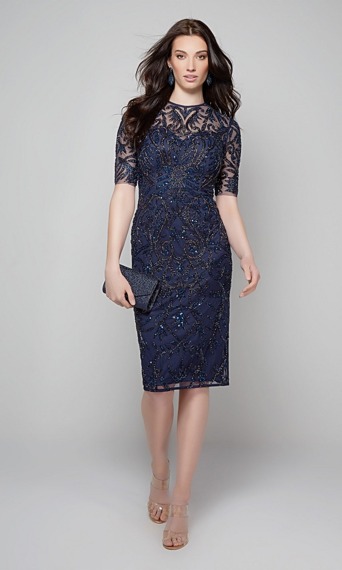 A woman in an Alyce 27583 midnight knee-length semi-formal dress with sheer sleeves, holding a matching clutch and wearing nude heels, stands against a light gray background.