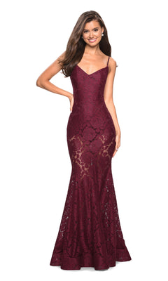 A woman stands smiling with one hand on her hip in a sleeveless, deep red Sheer Lace Mermaid Prom Dress: La Femme 27584 featuring thin straps and a fitted mermaid silhouette.