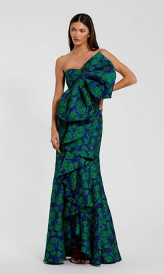 A woman wears the Mac Duggal 27593 long formal evening gown, featuring green floral brocade and dramatic one-shoulder ruffles, posed against a dark blue background.