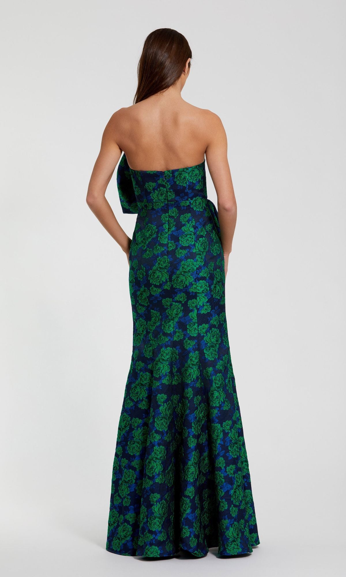 A woman with long brown hair wears the Mac Duggal 27593 Long Formal Evening Gown, a strapless dress with a blue and green floral pattern, standing with her back to the camera against a plain background.