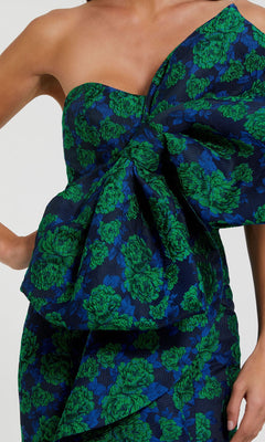A woman wears the Mac Duggal 27593 long formal evening gown—strapless, dark blue with a bold green floral print and an oversized ruffle on the bodice.