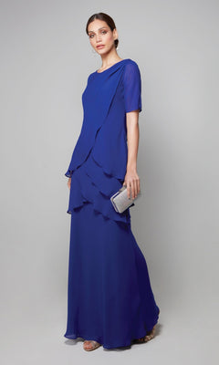 A woman in the Alyce 27593 long formal dress, featuring tiered layers of cobalt chiffon, holds a silver clutch while standing against a plain gray backdrop.