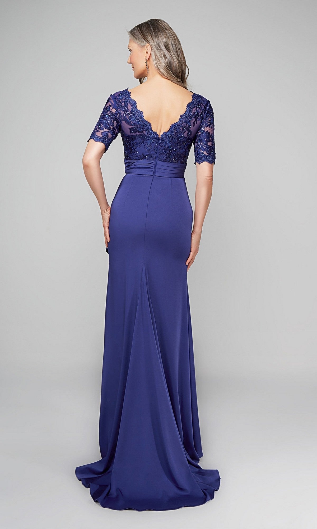A woman in the Alyce 27595 long formal dress—a floor-length, short-sleeve gown in deep blue with a lace bodice and elegant sheath skirt—faces away, glancing over her shoulder.