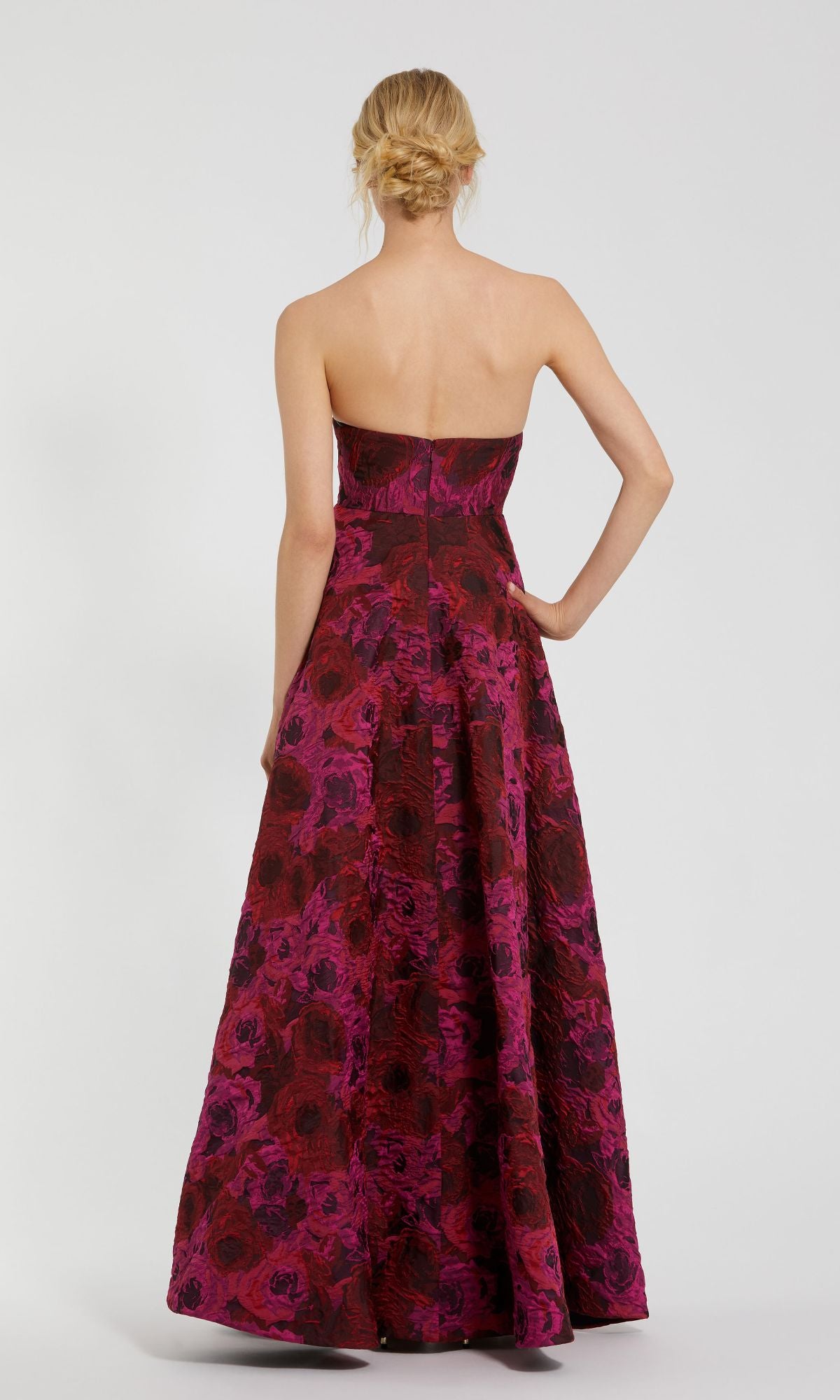 A woman faces away, wearing the Pink Floral Formal Evening Gown Mac Duggal 27596, a strapless brocade dress with a red and magenta floral pattern flowing down its floor-length silhouette.
