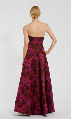 A woman faces away, wearing the Pink Floral Formal Evening Gown Mac Duggal 27596, a strapless brocade dress with a red and magenta floral pattern flowing down its floor-length silhouette.