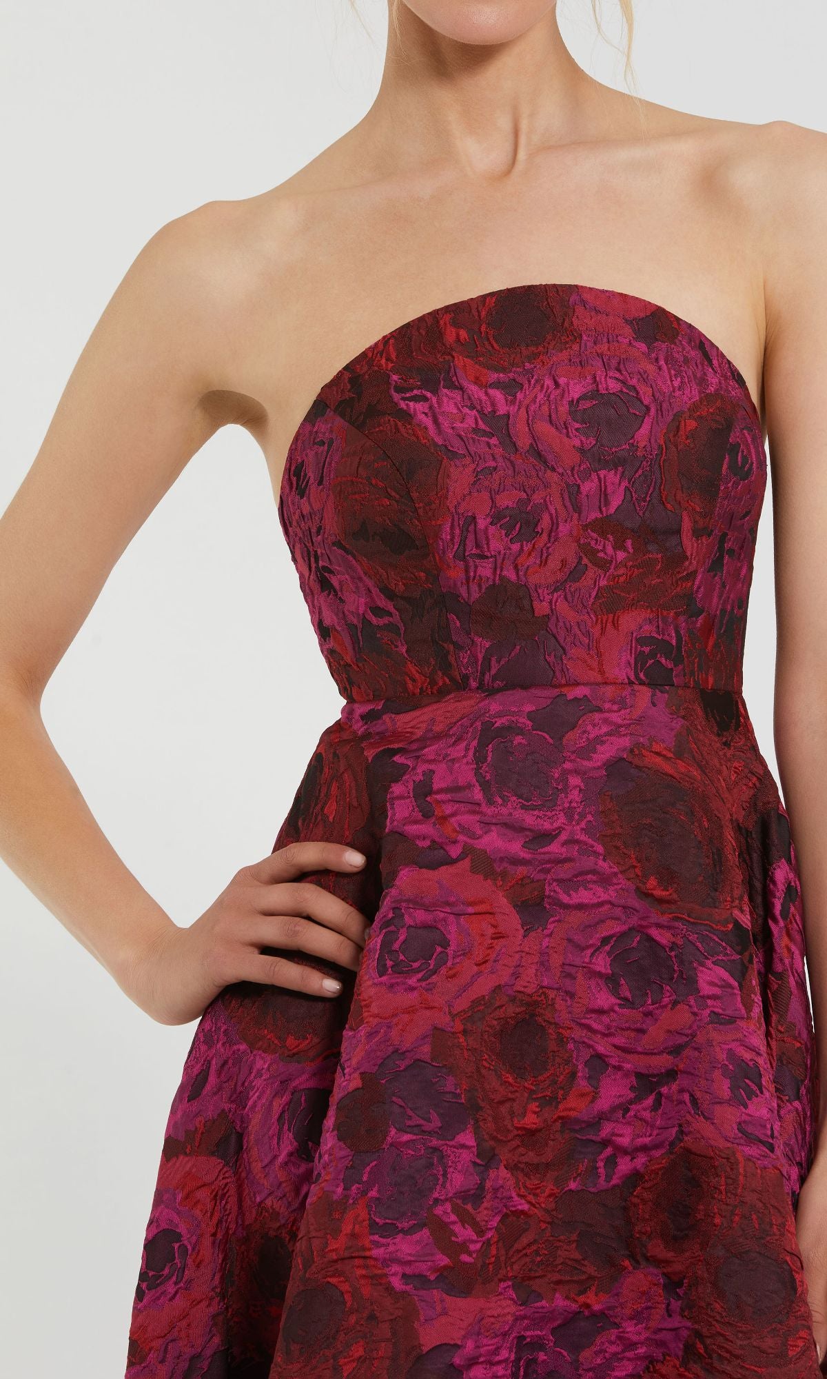 A woman poses with one hand on her hip, wearing the Mac Duggal 27596 Pink Floral Formal Evening Gown featuring a strapless brocade design with textured red and magenta floral patterns.