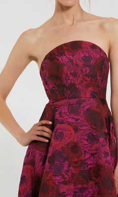 A woman poses with one hand on her hip, wearing the Mac Duggal 27596 Pink Floral Formal Evening Gown featuring a strapless brocade design with textured red and magenta floral patterns.