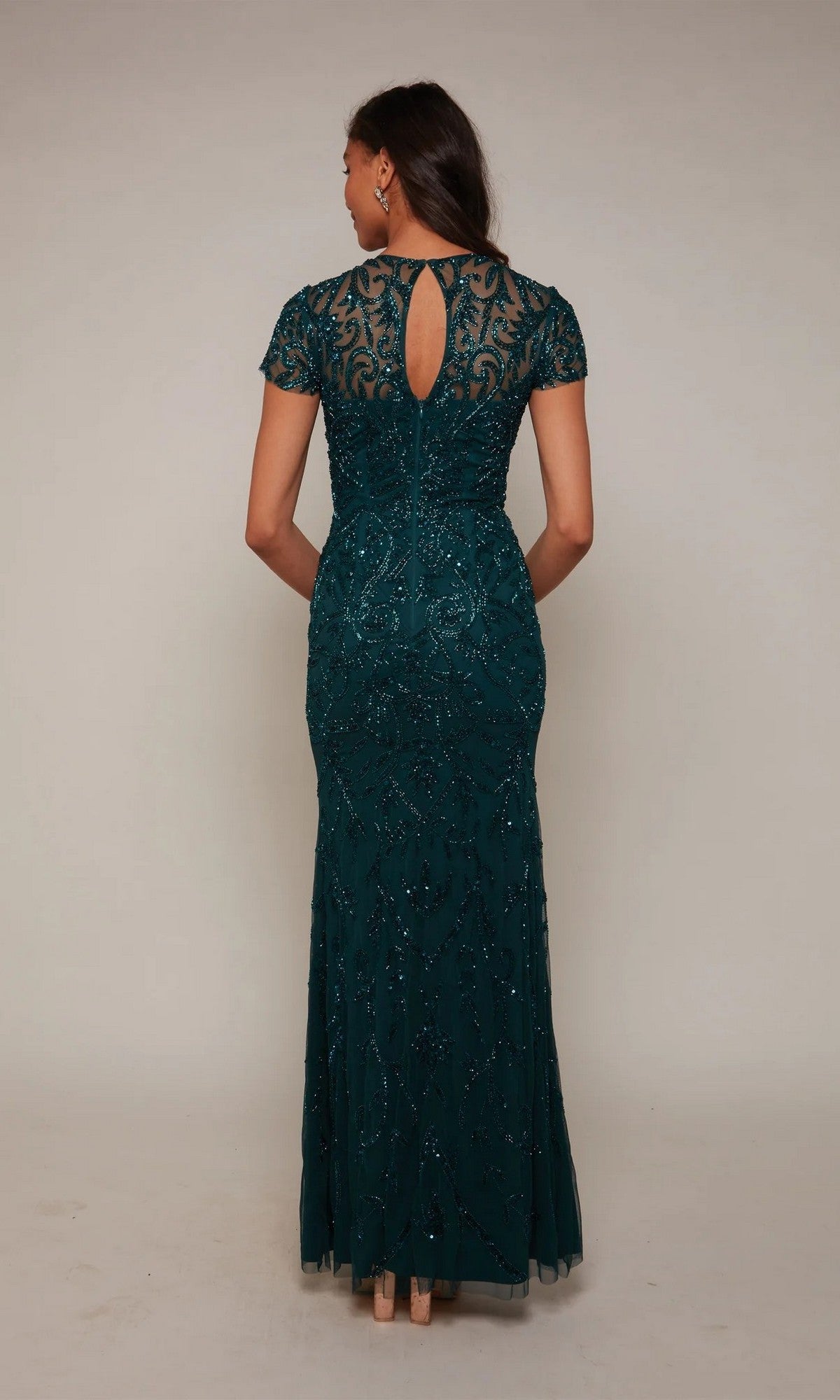 A woman poses in a studio wearing the Alyce 27607: a floor-length, short-sleeve, dark green beaded gown with intricate detailing and a keyhole back.