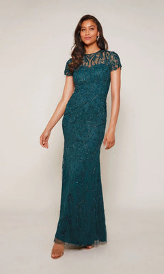 A woman stands against a neutral background wearing the Alyce 27607: a teal, floor-length formal dress featuring beads, short sleeves, and an illusion neckline.