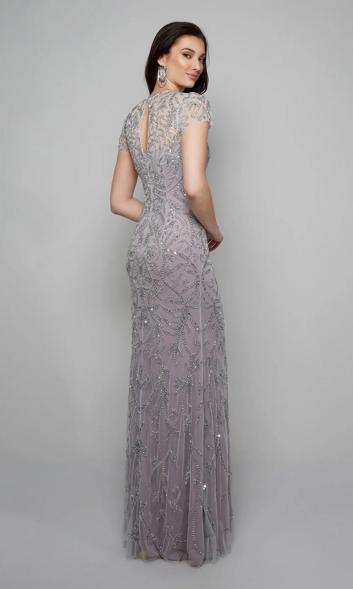A woman faces away, wearing the Alyce 27607 long formal dress in lilac. This floor-length gown features short sleeves, a keyhole back, and intricate beaded embellishments, all set against a plain gray background.