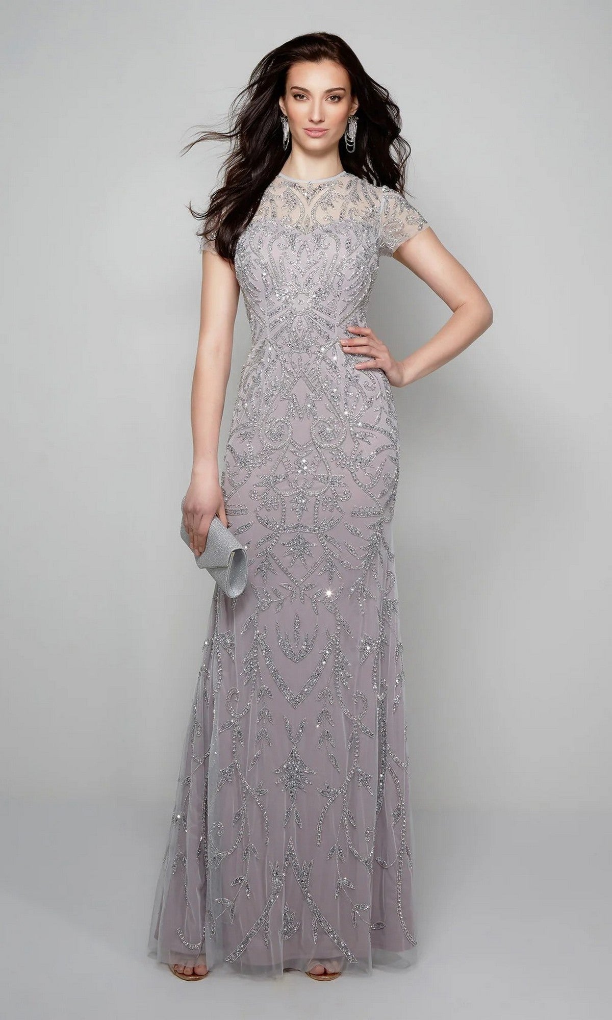 Woman wearing the Alyce 27607 long formal dress in light purple with silver beadwork, accessorized with a clutch and earrings, stands against a plain gray background.