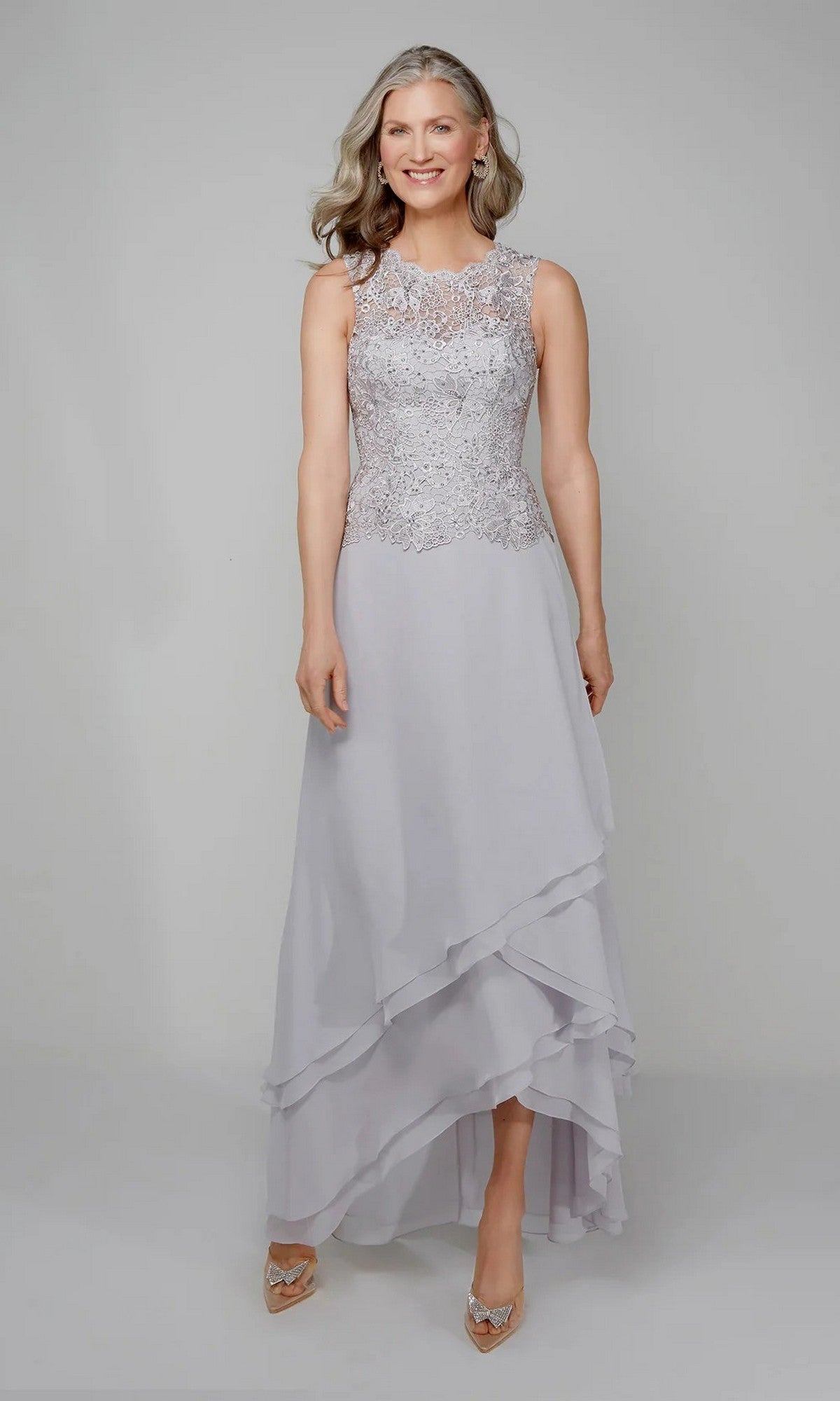 A woman in the Alyce 27608 long formal dress—sleeveless, light gray chiffon with a beaded lace bodice and layered asymmetrical skirt—stands against a plain background. An elegant choice for mothers of the bride.