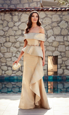 A woman in the Alyce 27612 long formal off-the-shoulder dress with a peplum waist and a gold clutch stands by a stone wall—a gorgeous mother-of-the-bride look.
