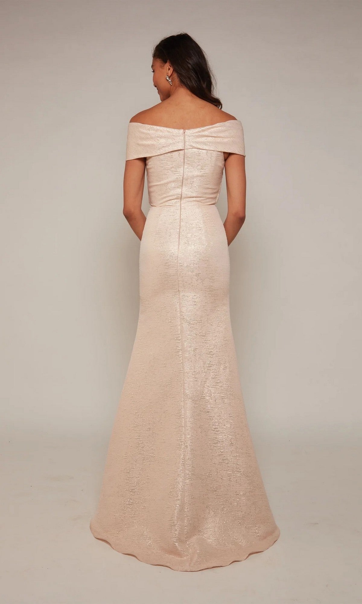 A woman stands with her back to the camera wearing the Alyce 27613, a floor-length light pink jacquard off-the-shoulder mother-of-the-bride dress, set against a plain studio backdrop.