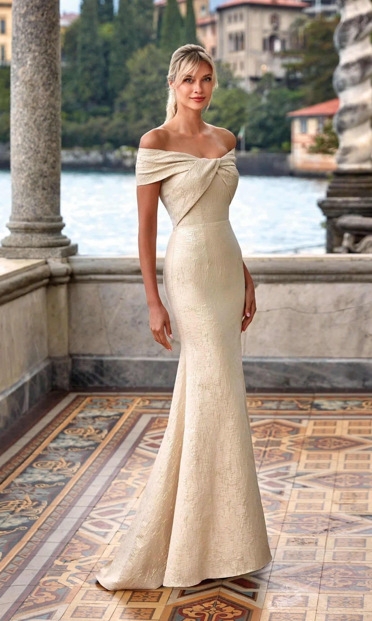 A woman stands on a tiled terrace with water and historic buildings behind her, wearing the Alyce 27613—a long, fitted, off-the-shoulder mother-of-the-bride dress in cream.