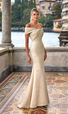 A woman stands on a tiled terrace with water and historic buildings behind her, wearing the Alyce 27613—a long, fitted, off-the-shoulder mother-of-the-bride dress in cream.
