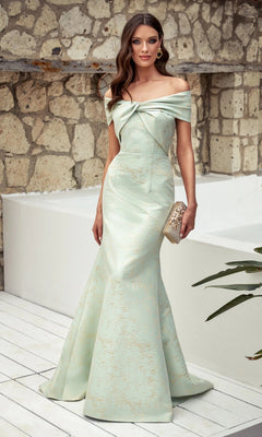 A woman in the Alyce 27613 long formal dress—a pale green off-the-shoulder gown with bow detail—holds a gold clutch while standing by a stone wall and white floor.