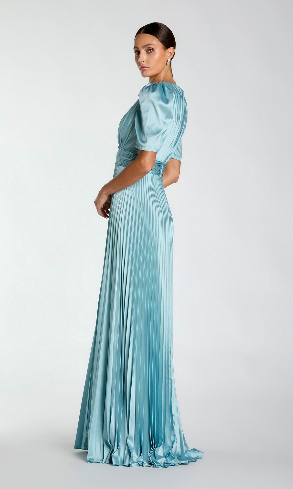 A woman poses sideways in the Mac Duggal 27616 long formal dress, featuring light blue pleats and short puff sleeves, exuding timeless elegance against a plain background.