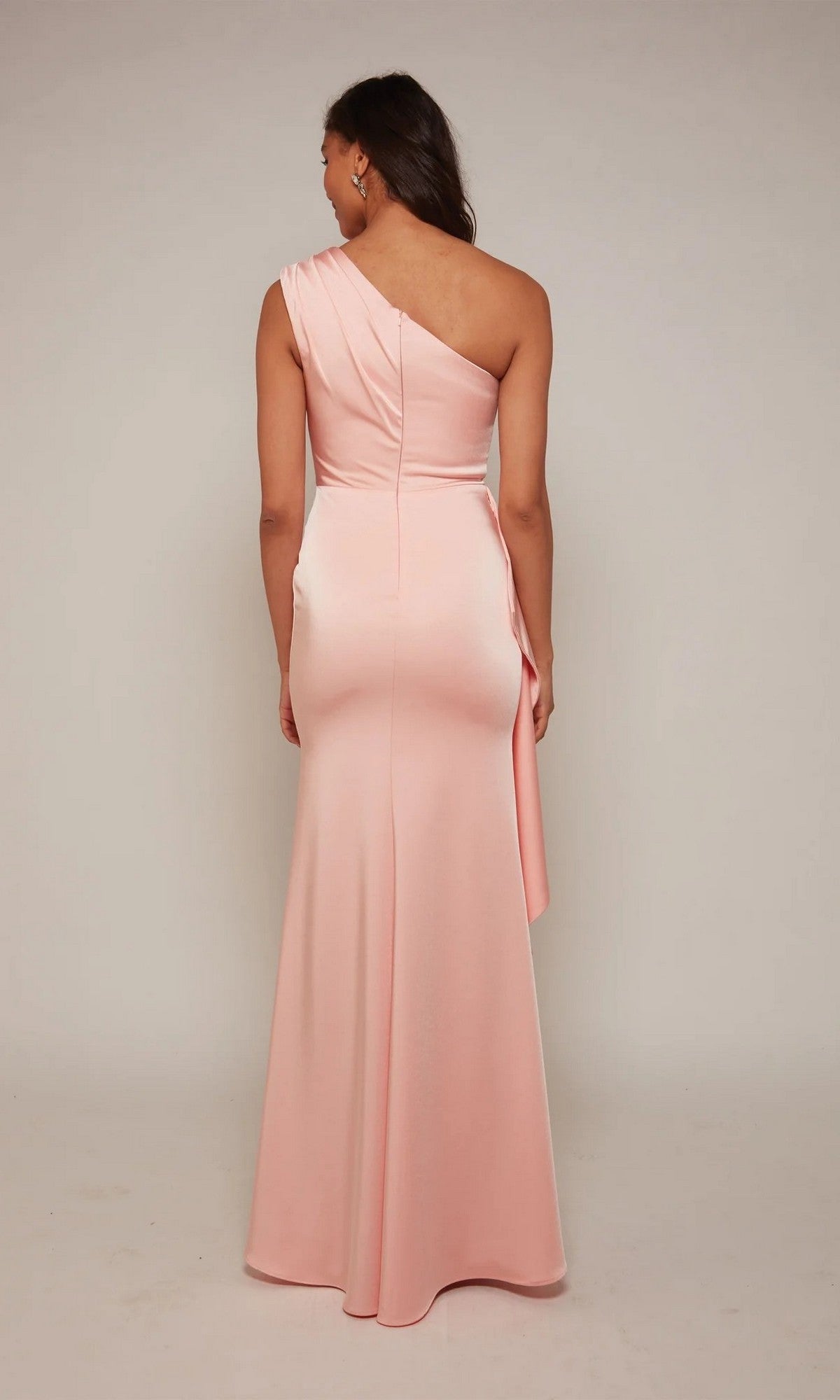 A woman stands facing away, wearing the Alyce 27624 long formal dress—an elegant one-shoulder gown in light pink with a fitted waist and flared skirt—against a plain background.