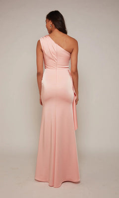 A woman stands facing away, wearing the Alyce 27624 long formal dress—an elegant one-shoulder gown in light pink with a fitted waist and flared skirt—against a plain background.
