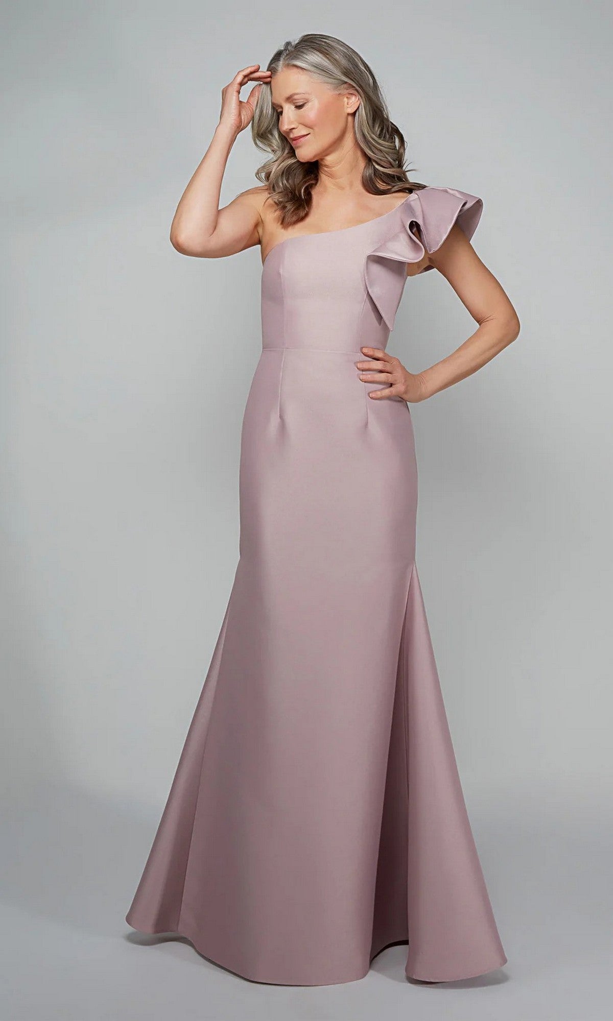 A woman poses with one hand on her head, wearing the Alyce 27641—a floor-length, light mauve one-shoulder formal dress with ruffled detail—an elegant choice for a mother-of-the-bride look against a plain background.