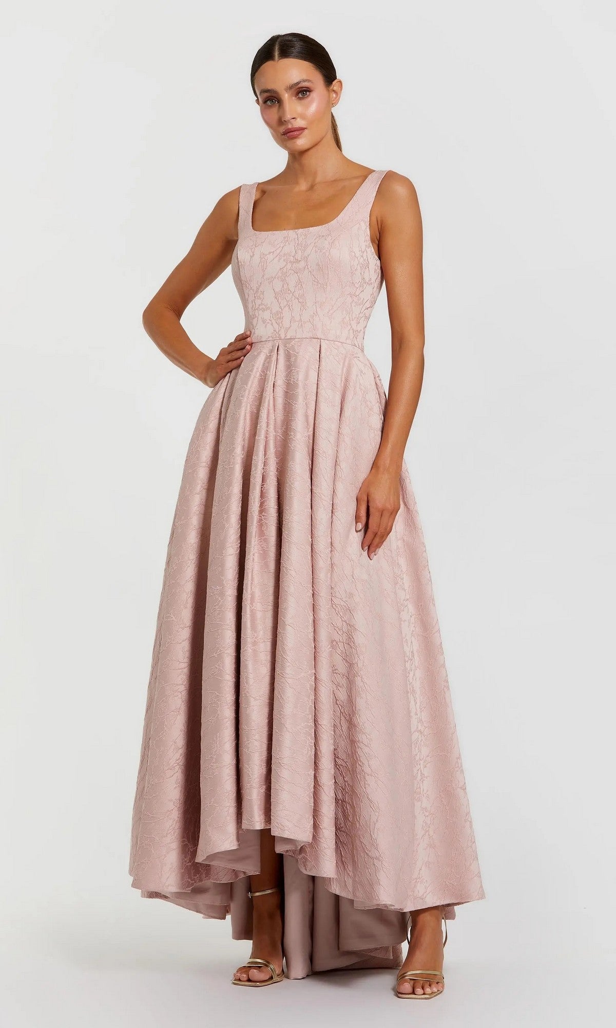 A woman stands against a plain white background wearing the Mac Duggal 27643 High-Low Formal Dress in blush, a sleeveless jacquard A-line dress with a textured high-low hem, styled with strappy heels.