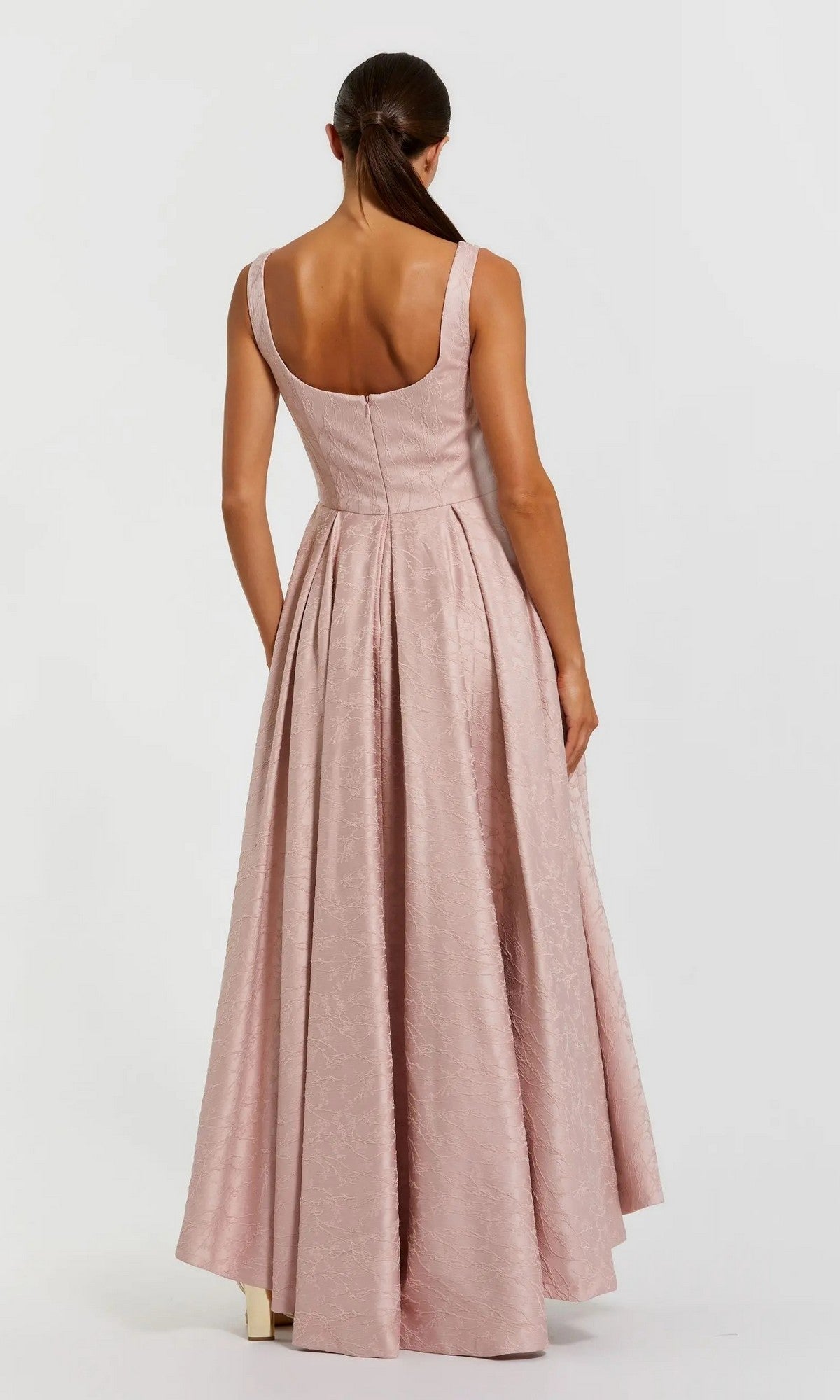 A woman with long brown hair styled in a low ponytail is seen from behind, standing against a plain light background and wearing the Mac Duggal 27643 High-Low Formal Dress—a sleeveless, floor-length gown in light pink.