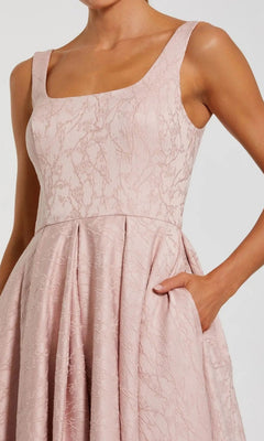 A woman wears the Mac Duggal 27643 High-Low Formal Dress, a sleeveless light pink jacquard A-line design featuring a fitted bodice, pleated skirt, and convenient dress pockets.