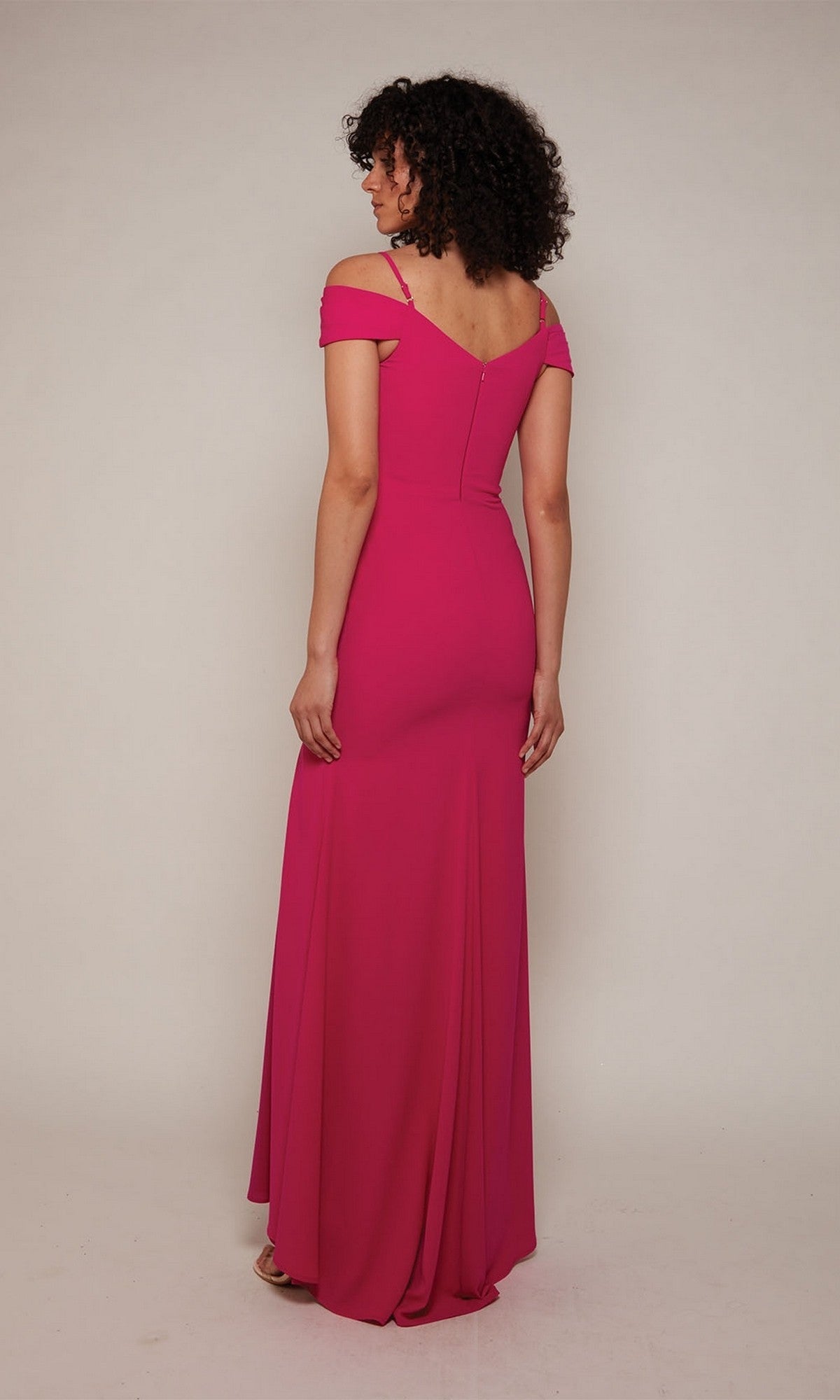 A woman stands in a studio, back to camera, wearing the Alyce 27645 long formal dress—a bright pink, floor-length gown featuring an off-the-shoulder neckline, fitted bodice with beaded embroidery, and a flowing skirt.