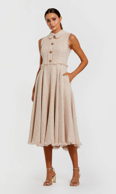 A woman models the Mac Duggal 27658 semi-formal midi dress, a sleeveless boucle design with large front buttons, one hand in her pocket and paired with strappy heels.