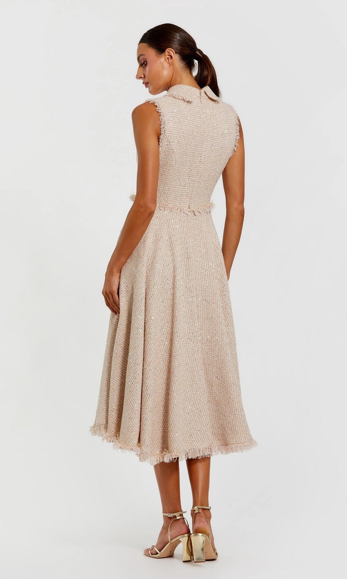 A woman stands facing away, wearing the Mac Duggal 27658 Semi-Formal Midi Dress—a sleeveless beige bouclé collared dress with a frayed hem—paired with strappy high-heeled sandals.