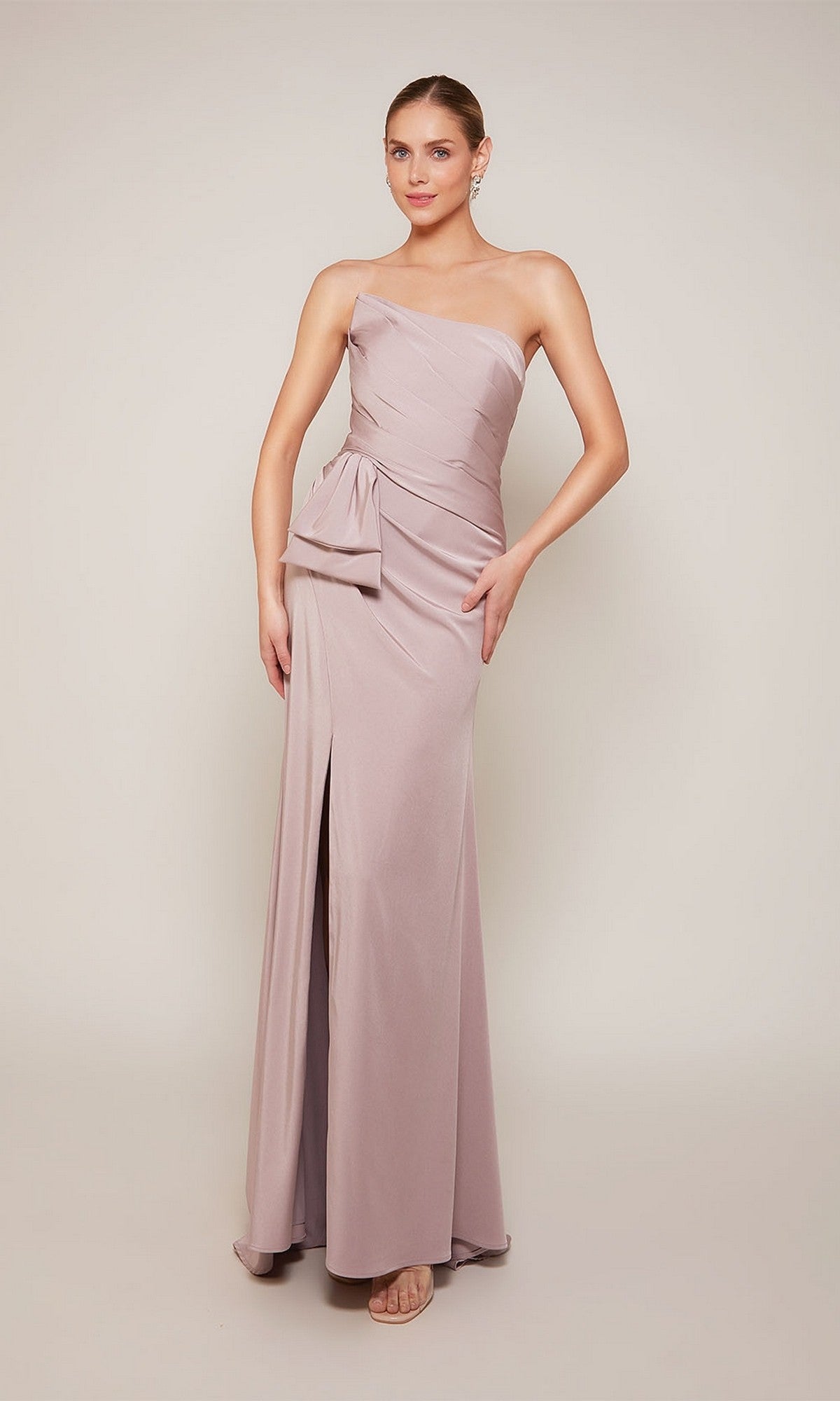 A woman wears the Alyce 27658 Long Formal Dress in rosewood—a one-shoulder, floor-length stretch crepe gown with a side slit and decorative bow—standing against a plain background.