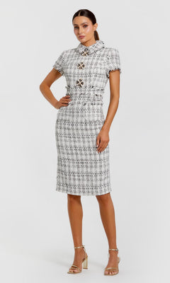 A woman poses against a plain white background wearing the Mac Duggal 27660 knee-length day dress in ivory, featuring a white and black plaid boucle pattern with jeweled buttons, paired with gold heels.
