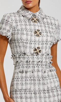 A woman wears the Mac Duggal 27660 knee-length day dress with short sleeves, a collared black and white plaid design, fringe details, and oversized jeweled buttons down the front.